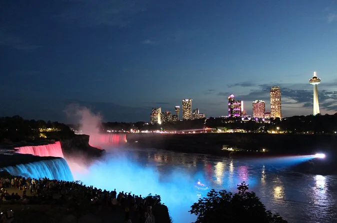 Niagara Falls lit in blue and pink at night with the skyline of Niagara Falls, Ontario, behind